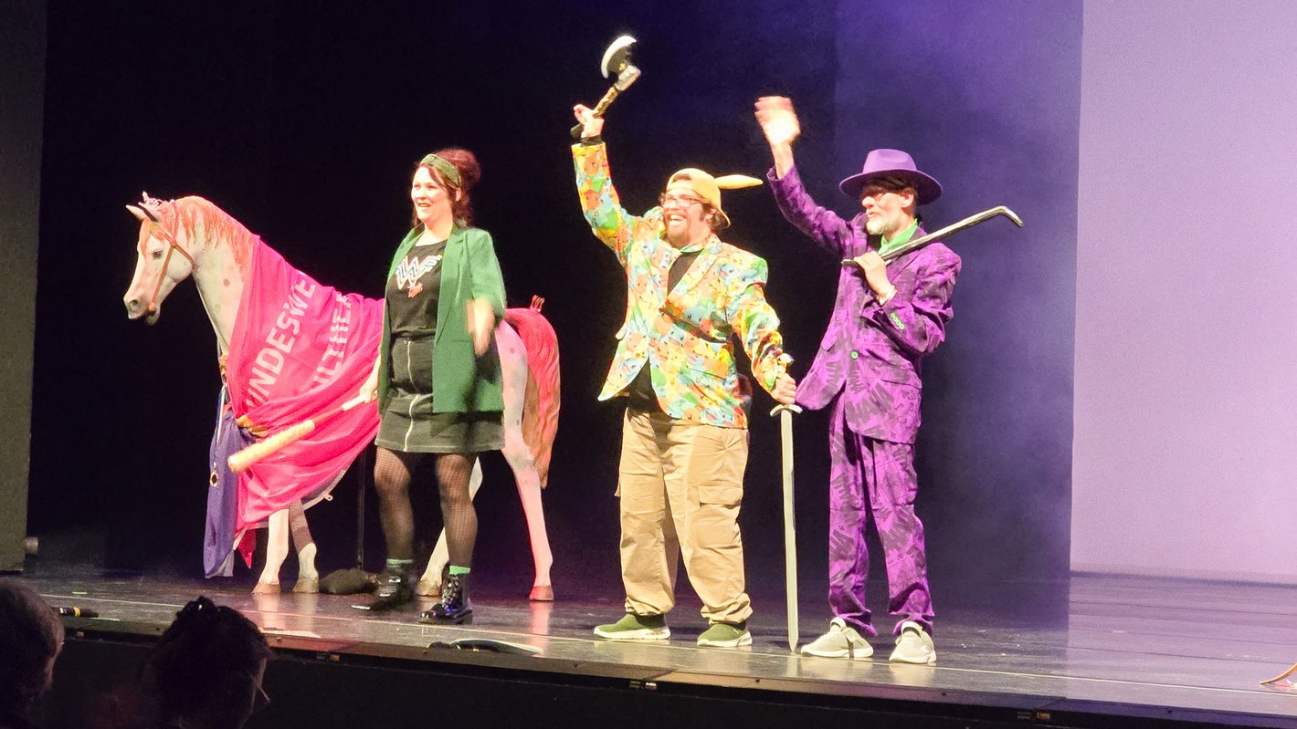 Drei Personen in bunter Kleidung stehen zusammen mit einer lebensgroßen Pferdefigur auf einer Theaterbühne und winken ins Publikum.