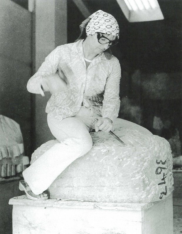 Eine Bildhauerin mit Schutzbrille und in weiß gekleidet sitzt auf einem schon behauenen Steinsockel und bearbeitet diesen.