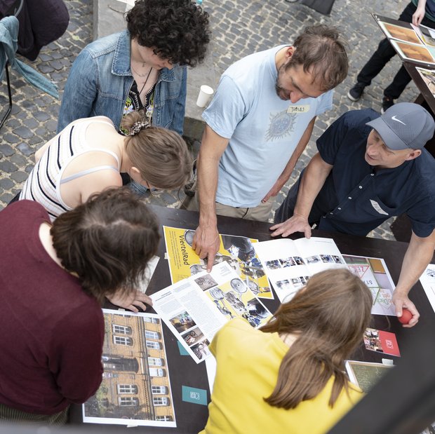 Personen die während des Projekt "Trans Urban X" um einen Tisch herum stehen und miteinander reden. Auf dem Tisch sind Broschüren und Fotos ausgebreitet.