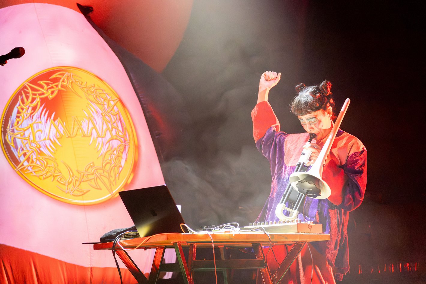 Eine Frau in bunter Kleidung steht an einem Tisch mit Musikgeräten und spielt Posaune.