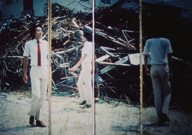 Ein Filmstill auf dem ein asiatisch aussehender Mann in weißer Kleidung und roter Krawatte vor einem Metall-Schrottberg entlang geht.