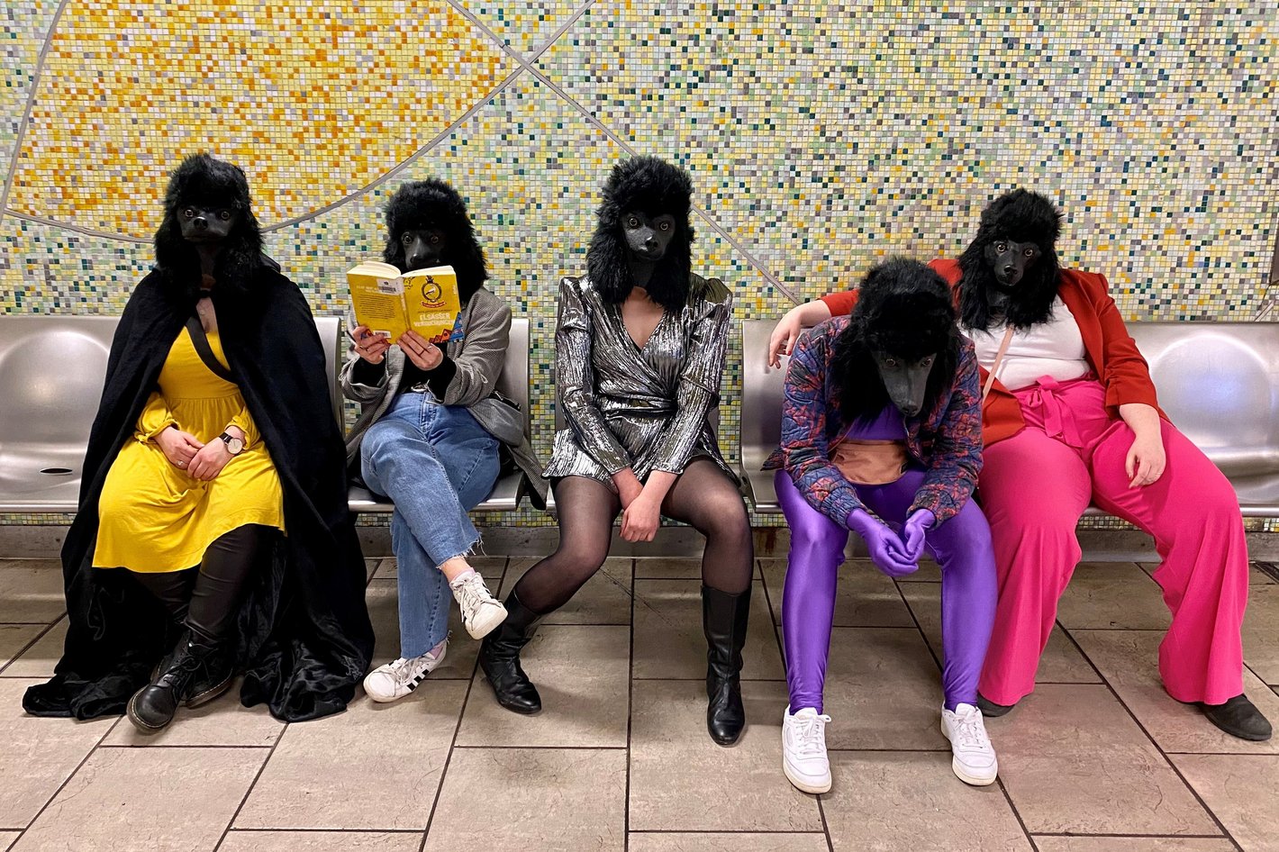 Fünf Personen mit Hundemasken und bunt gekleidet sitzen auf einer Bank in einer U-Bahnstation.